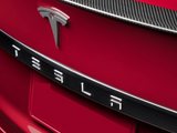 A red car features a prominent Tesla emblem and lettering on the rear, with a carbon-fiber trim above.