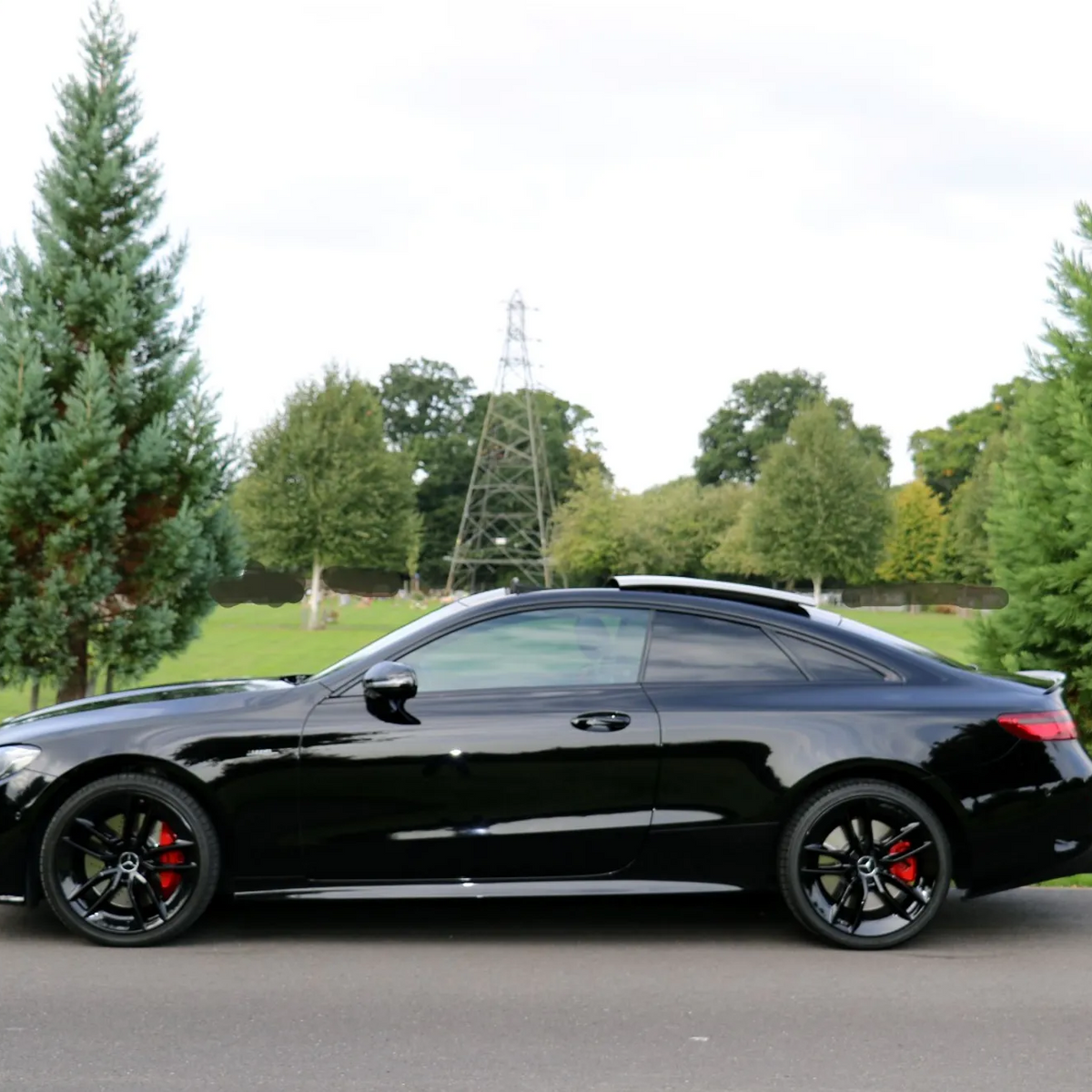 A sleek black car is parked on a paved road, surrounded by green grass and tall trees, under a partly cloudy sky.