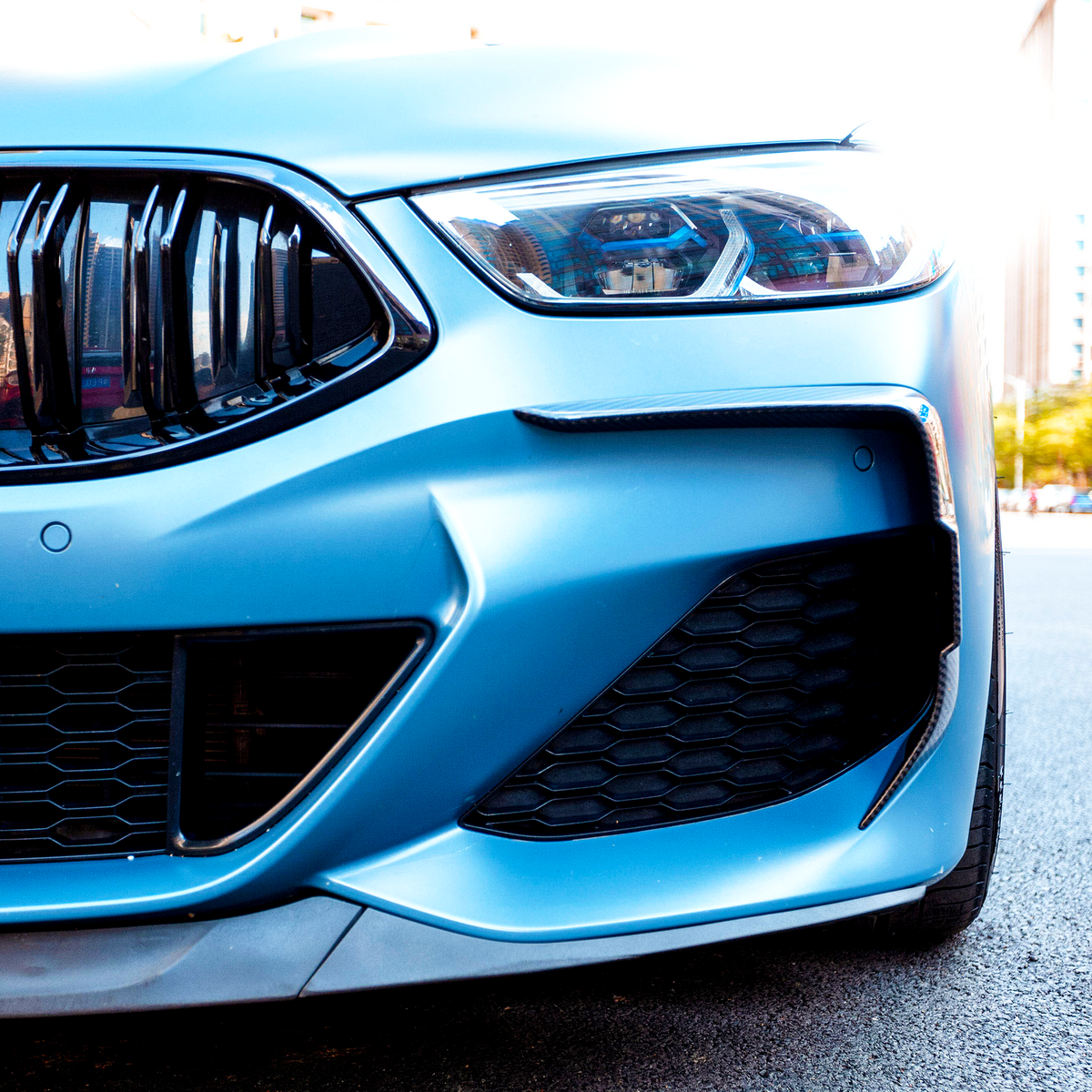 A sleek car grille and headlight on a blue vehicle, positioned on a city street with buildings and trees in the background.