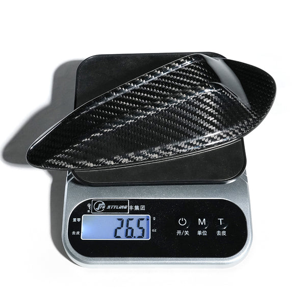 A carbon fiber car part rests on a digital scale, displaying a weight of 26.5 grams. Surrounding text includes "STI-LAN-9" and Chinese characters. The setting is plain, white.