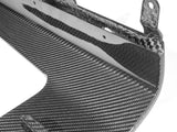 A close-up view of a carbon fiber automotive component with a textured pattern, featuring mounting brackets along the edge, set against a plain background.