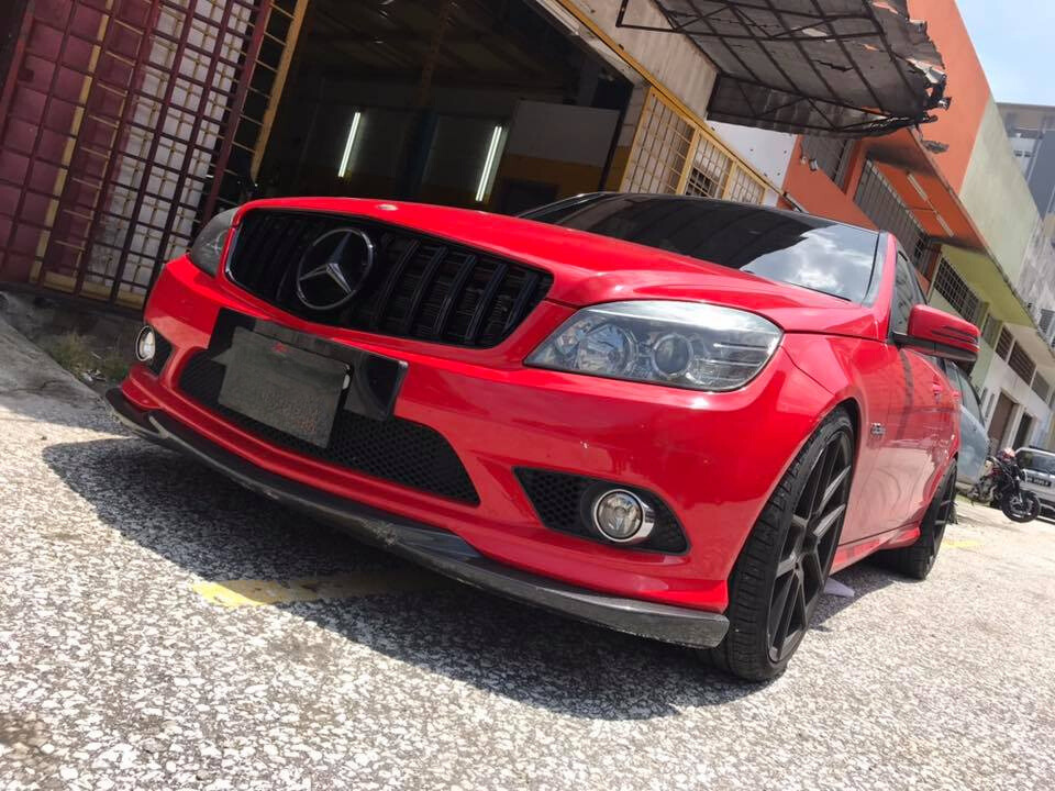 A red Mercedes-Benz car, parked with sleek, black wheels, sits in front of a building with an open garage door. The environment includes a textured pavement and nearby structures.