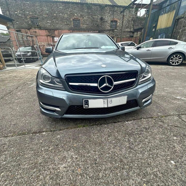 A silver Mercedes-Benz parked on a concrete surface, surrounded by several other vehicles, near a weathered building and wire fencing. License plates show symbols but no legible text.