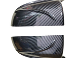 Two carbon fiber-patterned automotive mirror covers are positioned horizontally on a white background, showcasing their glossy finish and curved design, reflecting light and surroundings.