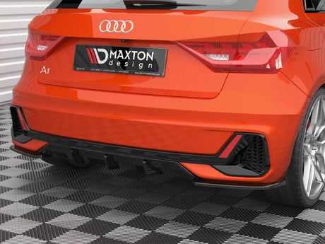 A bright orange Audi A1 is parked on a checkered floor, displaying a Maxton design rear spoiler and diffuser. The license plate reads "MAXTON design" with mentions of "SPLITTERS," "CAPSPOILERS," "DIFFUSERS," and "WWW.MAXTONDESIGN.COM."