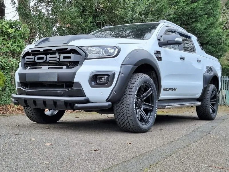 A white Ford pickup truck with "WILDTRAK" branding stands stationary on a paved surface. It features large tires and black detailing, surrounded by a leafy, wooded area.