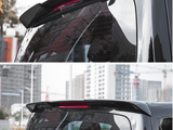 A black car rear spoiler with an integrated red brake light, attached above the rear window. The car is parked outdoors in a city area with buildings in the background.