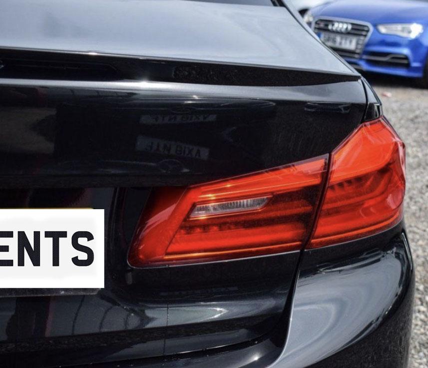 A close-up of a black car's rear with a vibrant red tail light. A license plate reads "ENTS." A blue car is parked nearby on a gravel surface.