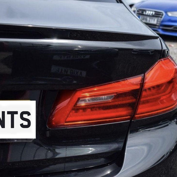 A close-up of a black car's rear with a vibrant red tail light. A license plate reads "ENTS." A blue car is parked nearby on a gravel surface.