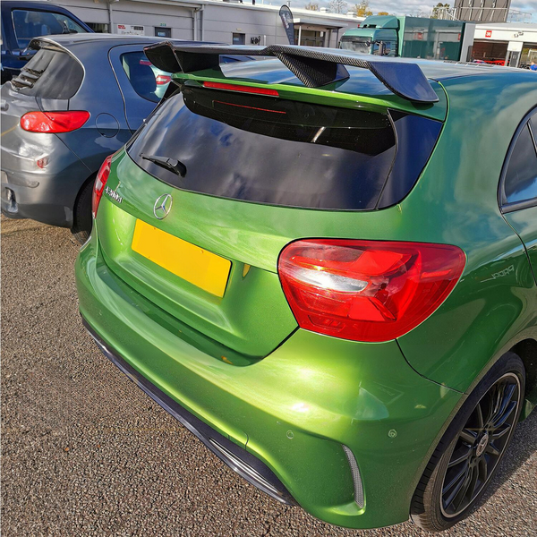 A green hatchback car, parked next to a gray car, features a prominent rear spoiler and a Mercedes logo. The setting is an outdoor parking area with buildings in the background.