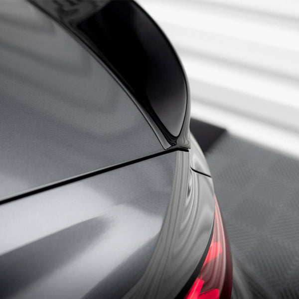 A shiny car's rear section with a sleek black spoiler, partially visible red taillights, is parked on a checkered pattern floor, set against a blurred, light-colored background.