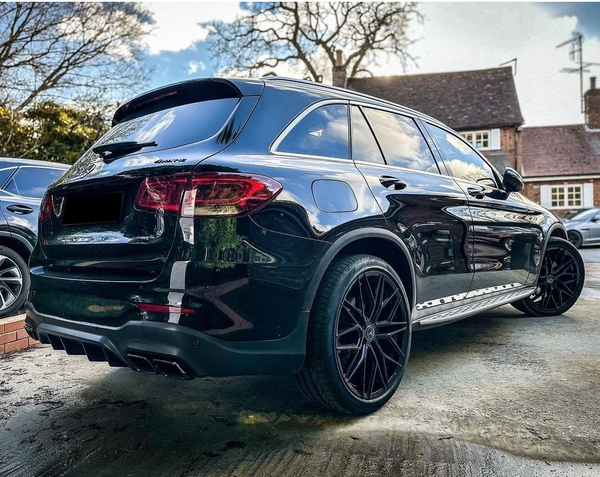 A black SUV with dark alloy wheels is parked on a driveway near brick houses under a blue sky. Text on the rear reads "4Matic." Trees and other cars are visible nearby.