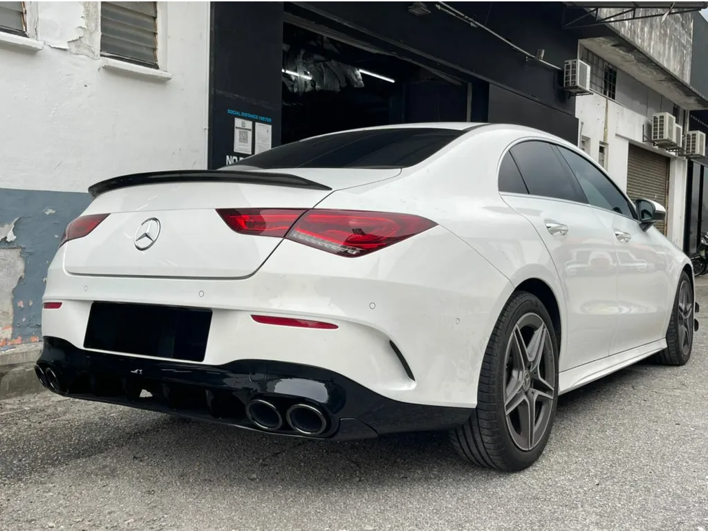A white sedan with a Mercedes emblem is parked beside a building, displaying sleek rear lights, tinted windows, and dual exhausts. The background includes nearby industrial structures.