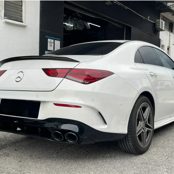A white sedan with a Mercedes emblem is parked beside a building, displaying sleek rear lights, tinted windows, and dual exhausts. The background includes nearby industrial structures.