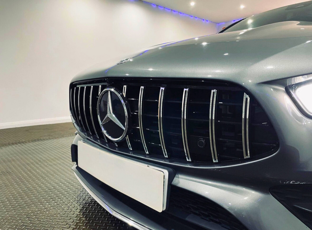 Car front with Mercedes-Benz logo on grille, parked in a lit indoor space with a metallic floor and white walls. A white license plate is attached below the grille.