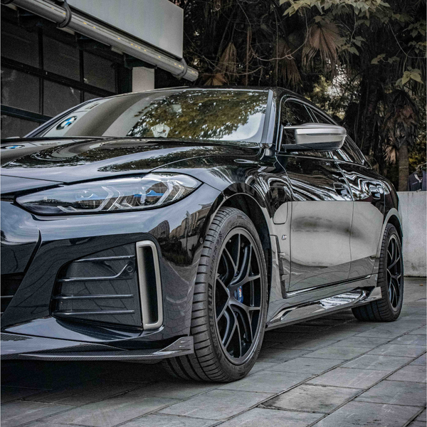 A sleek, black car is parked on a tiled pavement. The setting includes a modern building on the left and lush trees in the background, creating an urban yet natural ambiance.