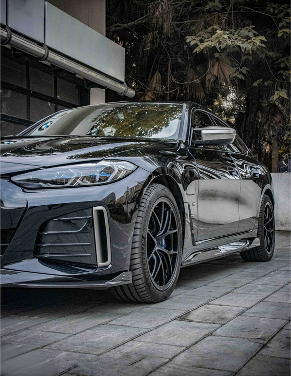 A sleek, black car is parked on a tiled pavement. The setting includes a modern building on the left and lush trees in the background, creating an urban yet natural ambiance.