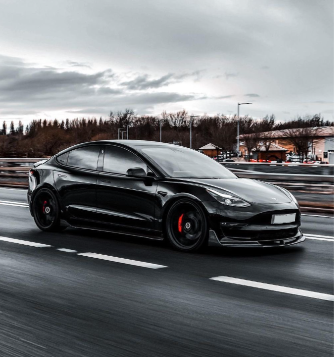 A black car with tinted windows drives quickly on a multi-lane road. The background features blurred trees and buildings under an overcast sky.