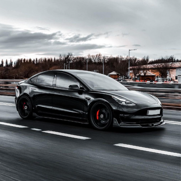 A black car with tinted windows drives quickly on a multi-lane road. The background features blurred trees and buildings under an overcast sky.