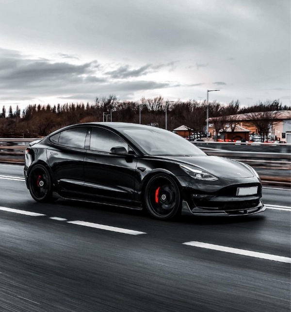 A black car with tinted windows drives quickly on a multi-lane road. The background features blurred trees and buildings under an overcast sky.