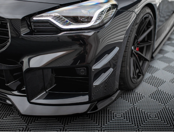 A sleek, black sports car shows its front headlight and grille, with a sporty bumper and aerodynamic features. It's parked on a textured, patterned surface, enhancing its modern appearance.