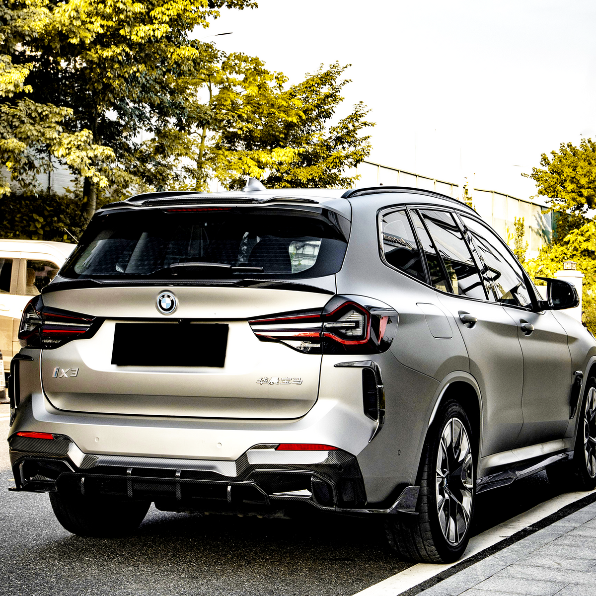A silver SUV is parked on a road beside a sidewalk, surrounded by green trees. The vehicle has a BMW logo and "iX3" and "Electric" badges visible.