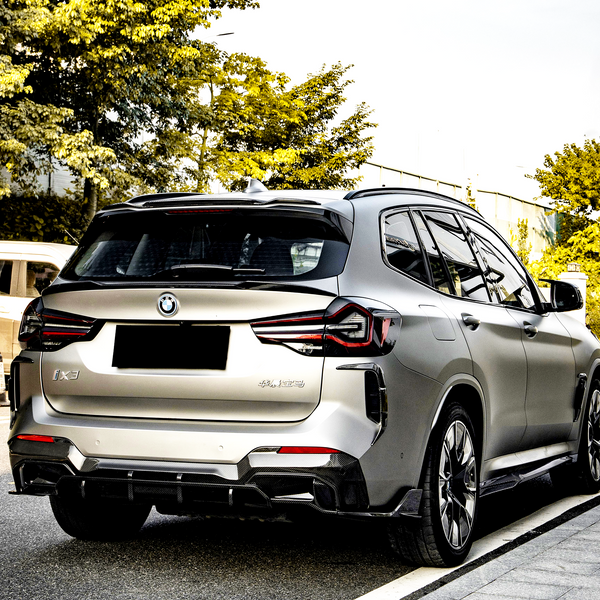 A silver SUV is parked on a road beside a sidewalk, surrounded by green trees. The vehicle has a BMW logo and "iX3" and "Electric" badges visible.