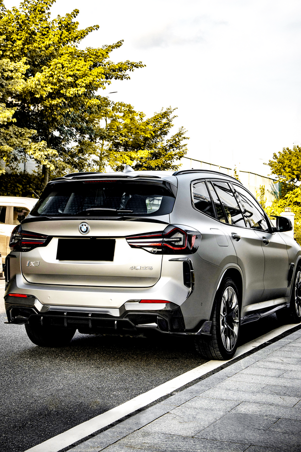 A silver SUV is parked on a road beside a sidewalk, surrounded by green trees. The vehicle has a BMW logo and "iX3" and "Electric" badges visible.