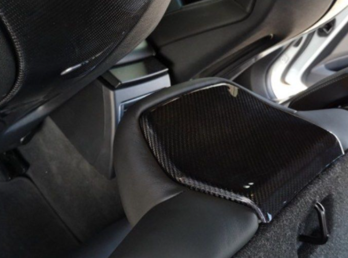 A black carbon fiber armrest is positioned inside a vehicle, with leather seating and a door slightly open in the background, revealing the car's interior environment.