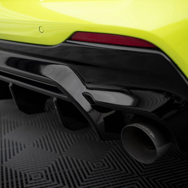 A bright yellow sports car's rear bumper features a glossy black trim and a visible exhaust pipe, set on a textured, geometric-patterned floor.