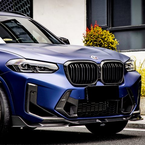 A blue SUV with prominent grille and headlights parked near a building, surrounded by leafy shrubs.