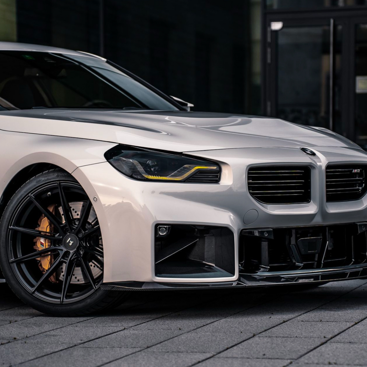 A silver car with black rims is parked on a paved surface. Its sleek design features modern headlights and a prominent grille, set against a dark urban backdrop.