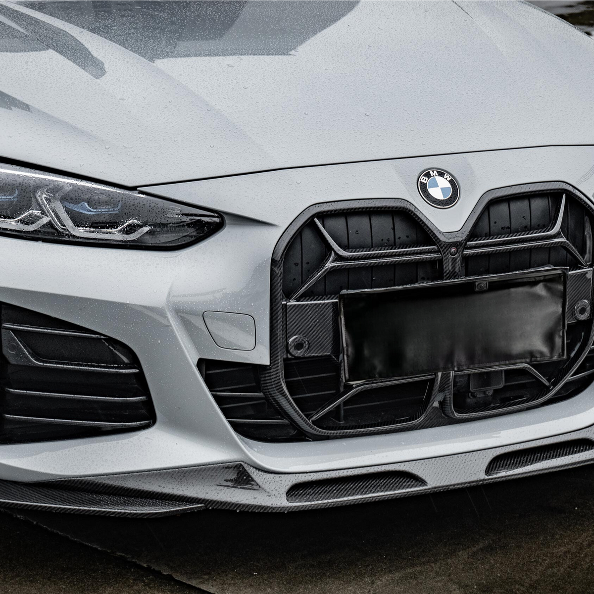 A matte gray BMW car with prominent grille and sleek headlights is parked on a wet, reflective ground surface, surrounded by outlined parking spaces.