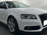 A white Audi car is parked indoors on a dark, reflective floor. It has black details, a prominent front grille, and a sleek, sporty design. The background is softly lit.