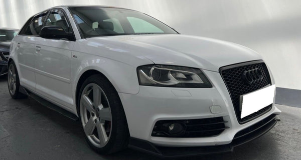 A white Audi car is parked indoors on a dark, reflective floor. It has black details, a prominent front grille, and a sleek, sporty design. The background is softly lit.
