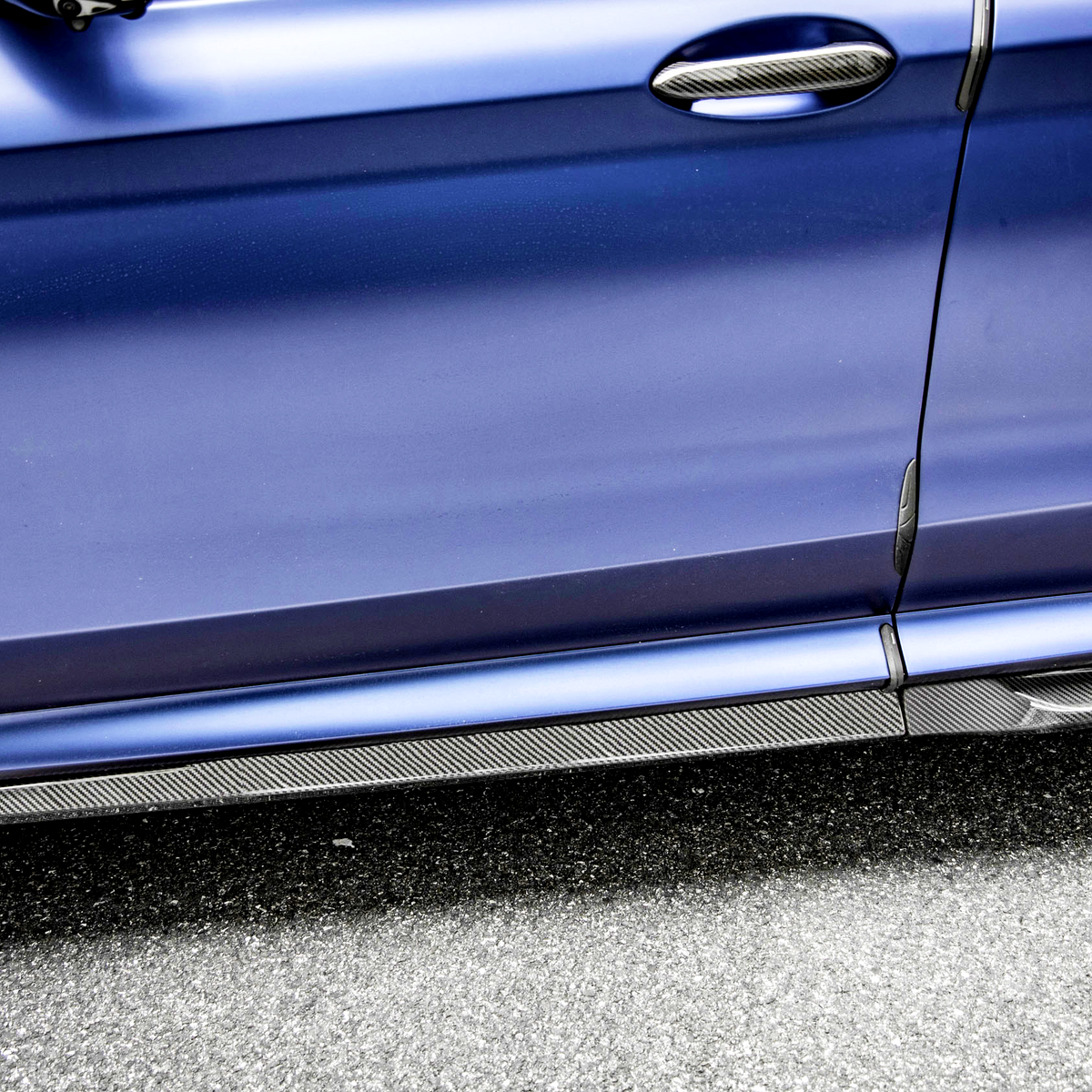 A blue car door with a carbon fiber handle and trim features an "X3 M" badge, parked on a textured gray asphalt surface.