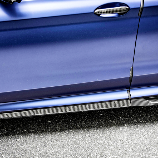 A blue car door with a carbon fiber handle and trim features an "X3 M" badge, parked on a textured gray asphalt surface.