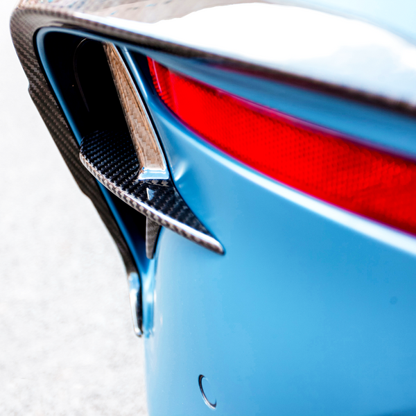 A car's rear end in close-up shows a carbon fiber vent, blue body, and red taillight. The scene is set on a light, neutral background.