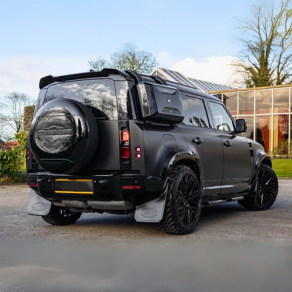 Black SUV parked on a road, equipped with a spare tire cover labeled "DEFENDER." It features large wheels and mud flaps, surrounded by a glass building and trees.