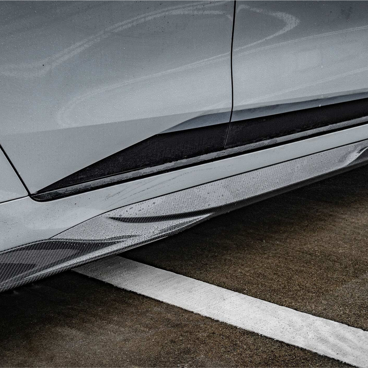 Car side skirt with "M PERFORMANCE" text is enhanced by raindrops. It's part of a sleek vehicle parked on wet pavement with a white painted line.