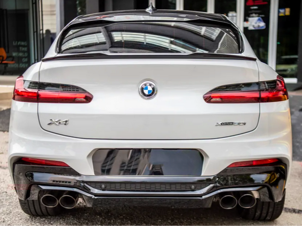 A white BMW X4 car is parked, displaying its rear view with distinctive taillights and dual exhaust pipes; it's positioned in an urban environment near a building entrance.