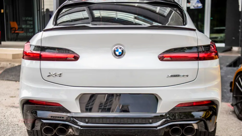 A white BMW X4 car is parked, displaying its rear view with distinctive taillights and dual exhaust pipes; it's positioned in an urban environment near a building entrance.