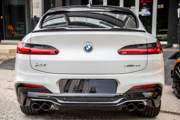 A white BMW X4 car is parked, displaying its rear view with distinctive taillights and dual exhaust pipes; it's positioned in an urban environment near a building entrance.
