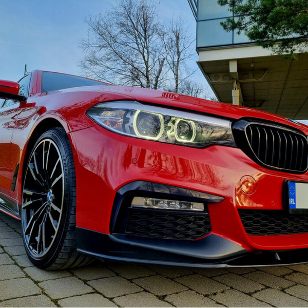 Red car parked on brick pavement, featuring sleek black rims and distinctive front grille; modern building and bare trees in background. License plate reads "IRL."
