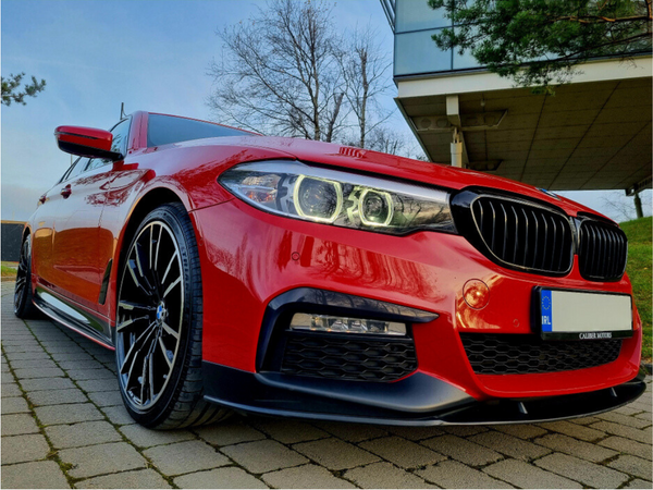 Red car parked on brick pavement, featuring sleek black rims and distinctive front grille; modern building and bare trees in background. License plate reads "IRL."