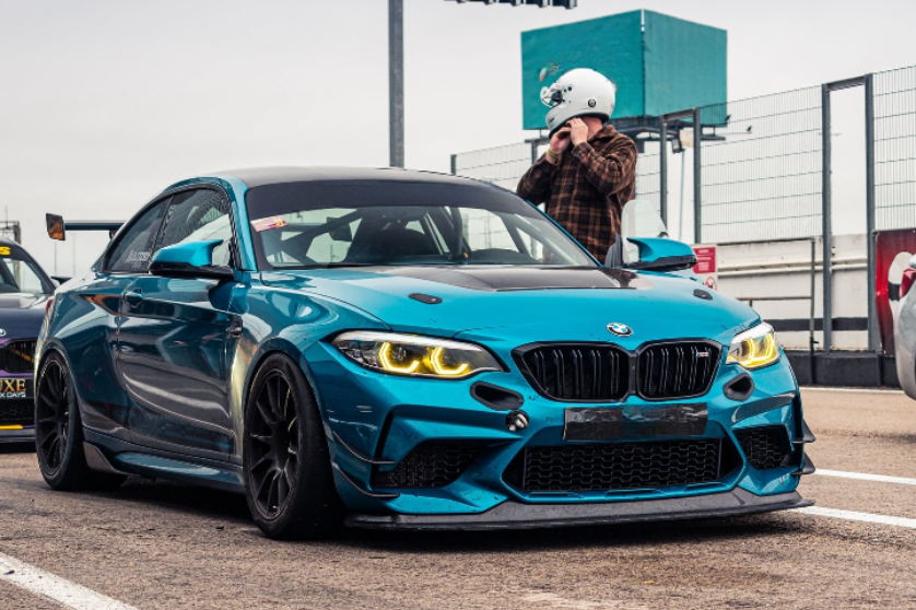 A blue sports car with aggressive styling is parked on a racetrack. Its headlights are illuminated. Nearby, a person wearing a helmet stands, adjusting the strap in a fenced area.