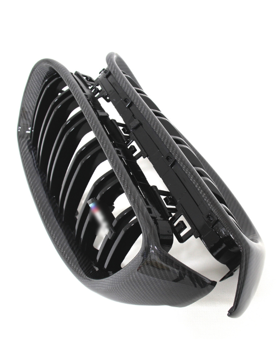 A black carbon fiber car grille with multiple vertical slats lies against a white background, displaying its glossy finish and intricate design details.