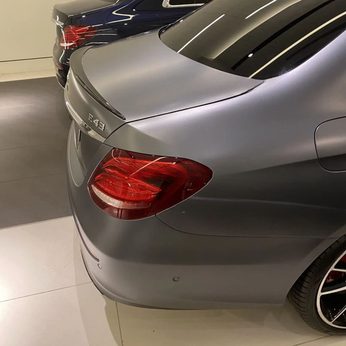 A gray luxury car's rear with "E 43" badge; parked beside another blue car inside a showroom with reflective flooring.