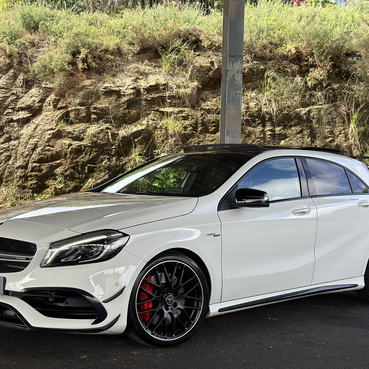 A white Mercedes-Benz car with AMG styling is parked under a structure. It is positioned on a dark paved surface, with a rocky, grassy hill in the background.