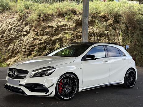 A white Mercedes-Benz car with AMG styling is parked under a structure. It is positioned on a dark paved surface, with a rocky, grassy hill in the background.
